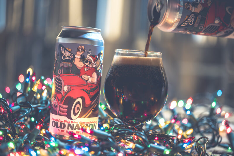 A can of Old Nation Frandor Claus beer in an aluminum can with a drawing of a Santa character with images from the city of Lansing and the beer name on the can label. surrounded by holiday lights. A beer glass is next to it with a dark beer being poured into it.