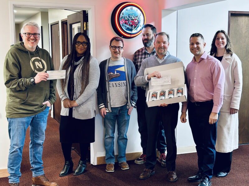 A group of people standing in front of a wall. One is holding a check up they are presenting to another person/